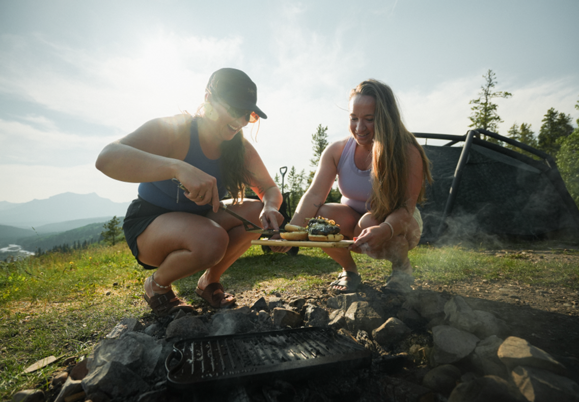 Two friends preparing burgers beside a campsite grill with mountains in the background.
