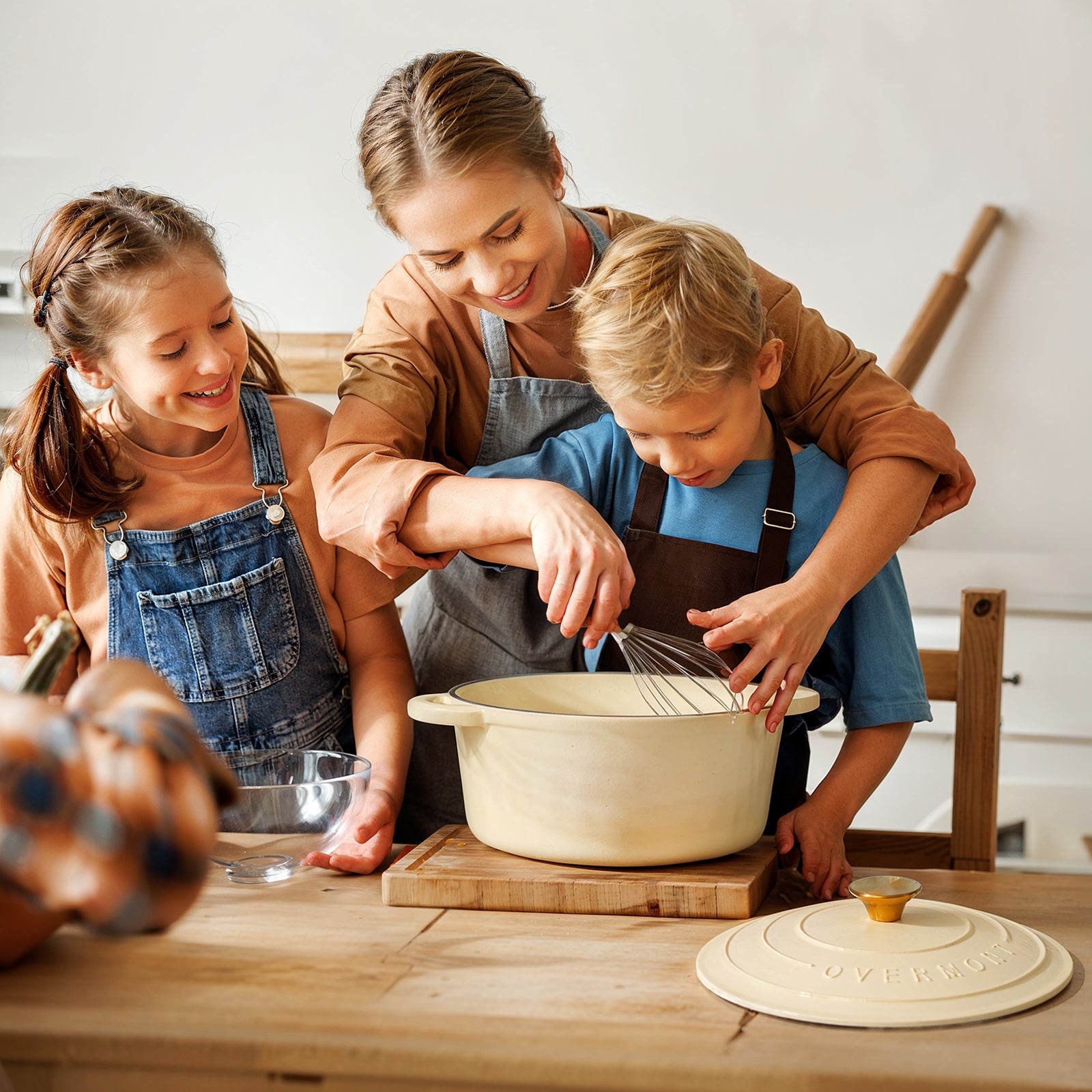 A woman and two children are cooking together in the kitchen using an cream white  enamel pot.